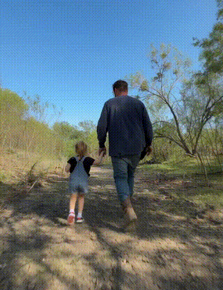 An adult and a young child walking together on a dirt path through trees on a sunny day.